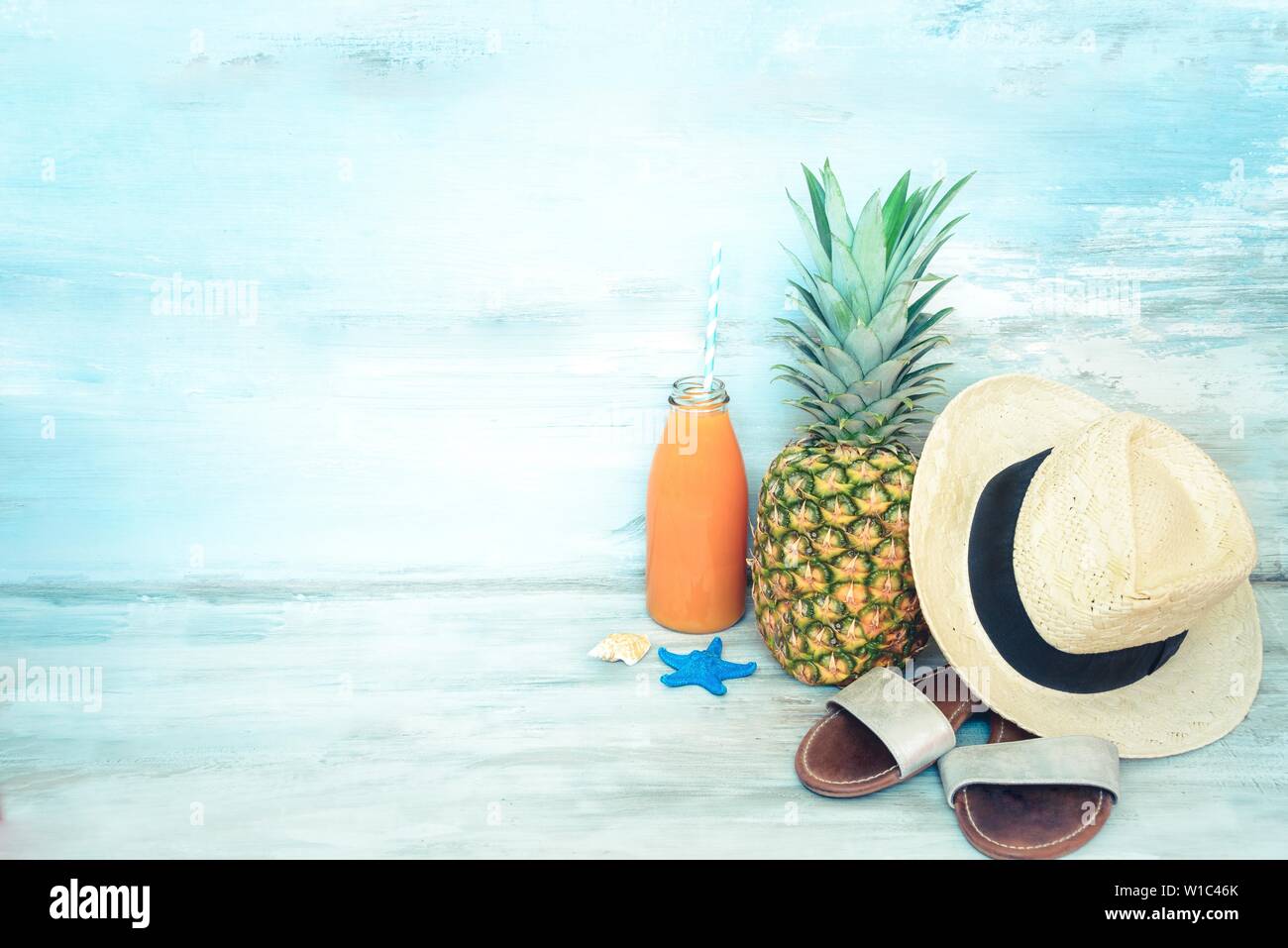 Summer concept stillife - ripe pineapple, straw hat, flip-flops and a bottle of multivitamin juice in front of a blue rustic wooden background. Stock Photo