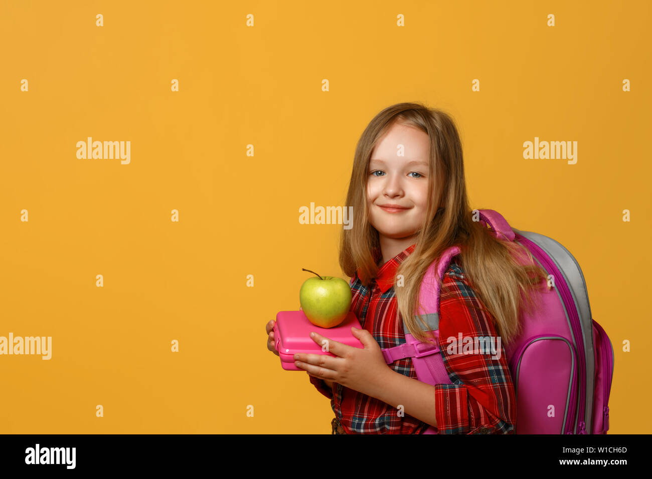 Portrait of a little girl schoolgirl on a yellow background. The child has a satchel behind his back and holds a lunchbox in his hands. Back to school Stock Photo