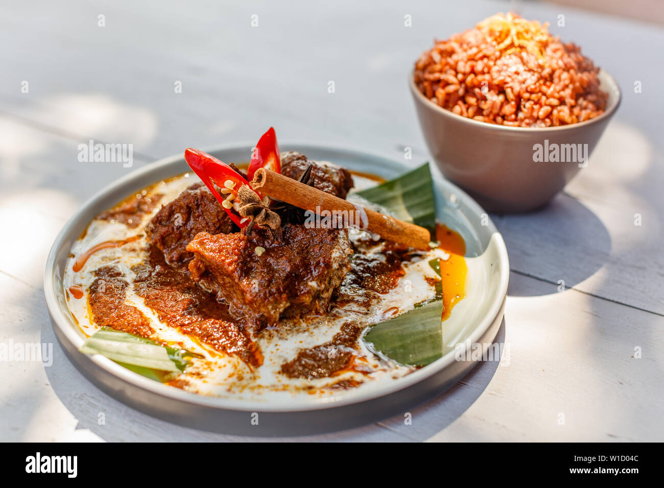 Beef Rendang on a plate with banana leaf, decorated with red chili and cinnamon stick. Served with nasi merah, steamed red rice. Indonesian cuisine. Stock Photo