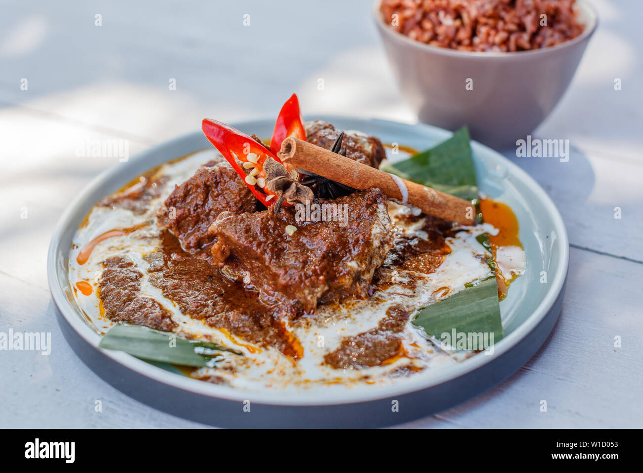 Beef Rendang on a plate with banana leaf, decorated with red chili and cinnamon stick. Served with nasi merah, steamed red rice. Indonesian cuisine. Stock Photo