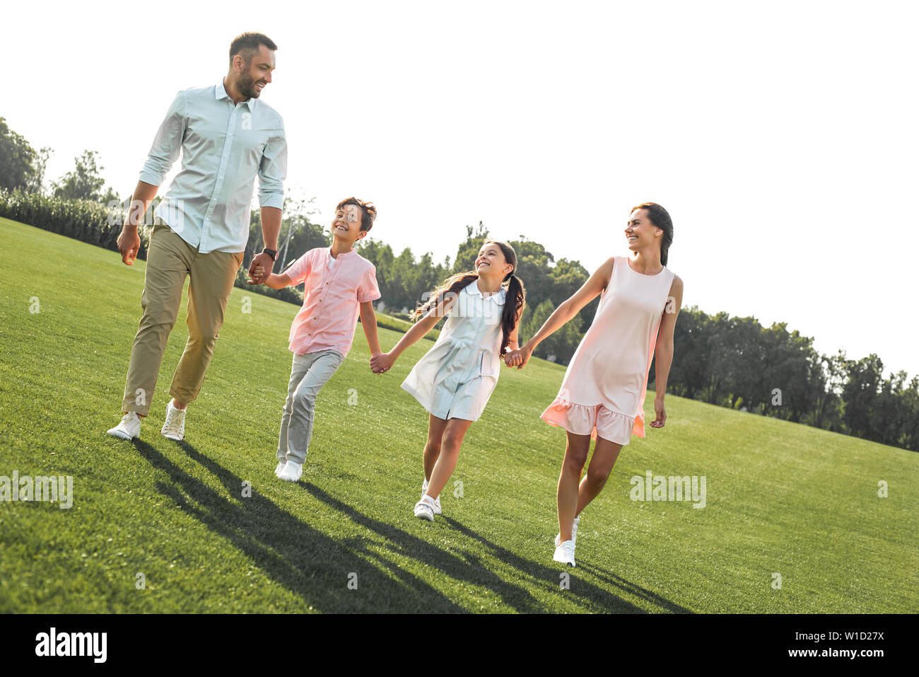 We are happy together Full length of positive and young family of four holding hands, smiling and walking outdoors . Picnic. Weekend Stock Photo