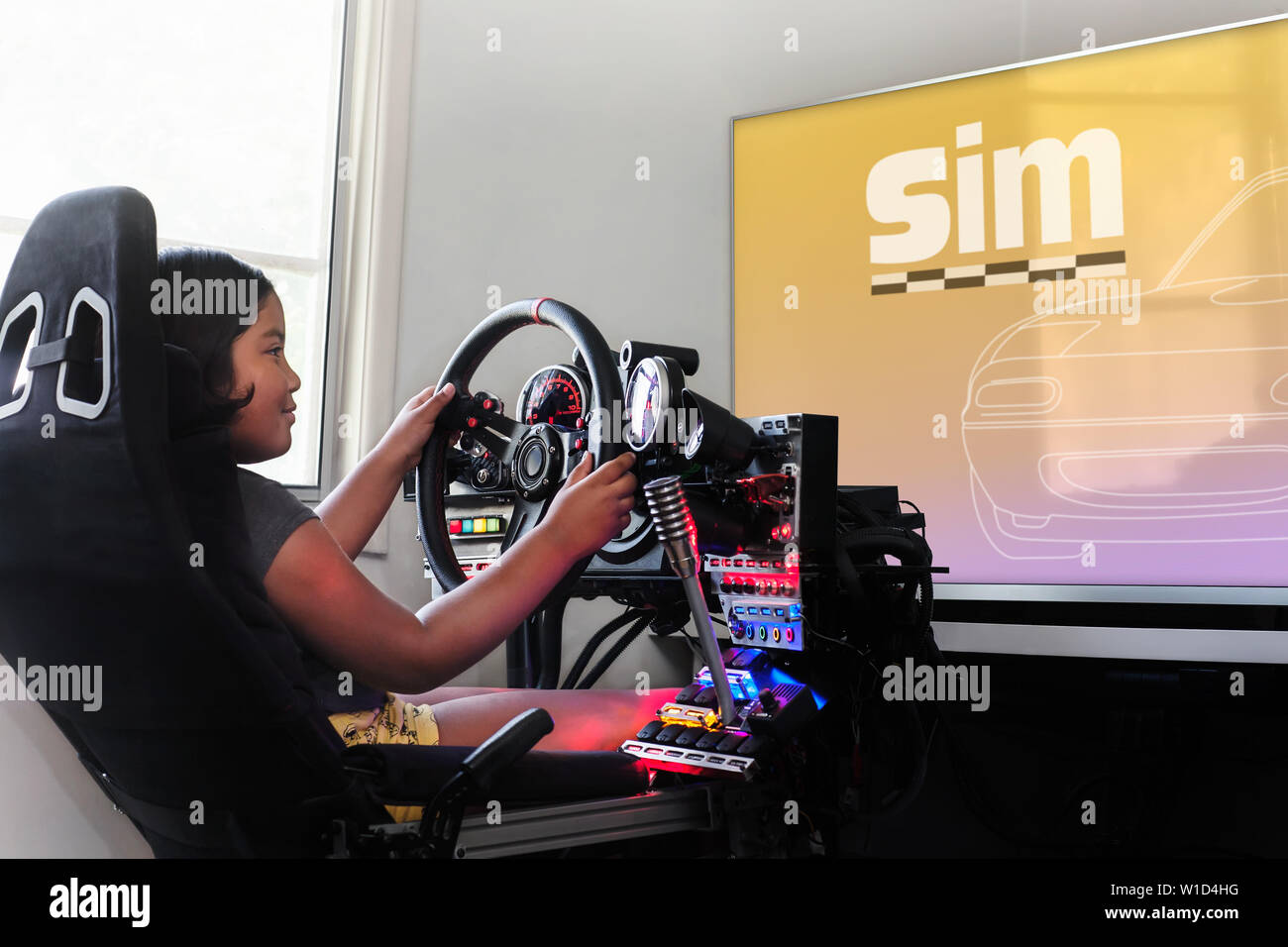 A little girl sitting in a gaming cockpit and holding the race steering wheel to play a racing simulation. Stock Photo