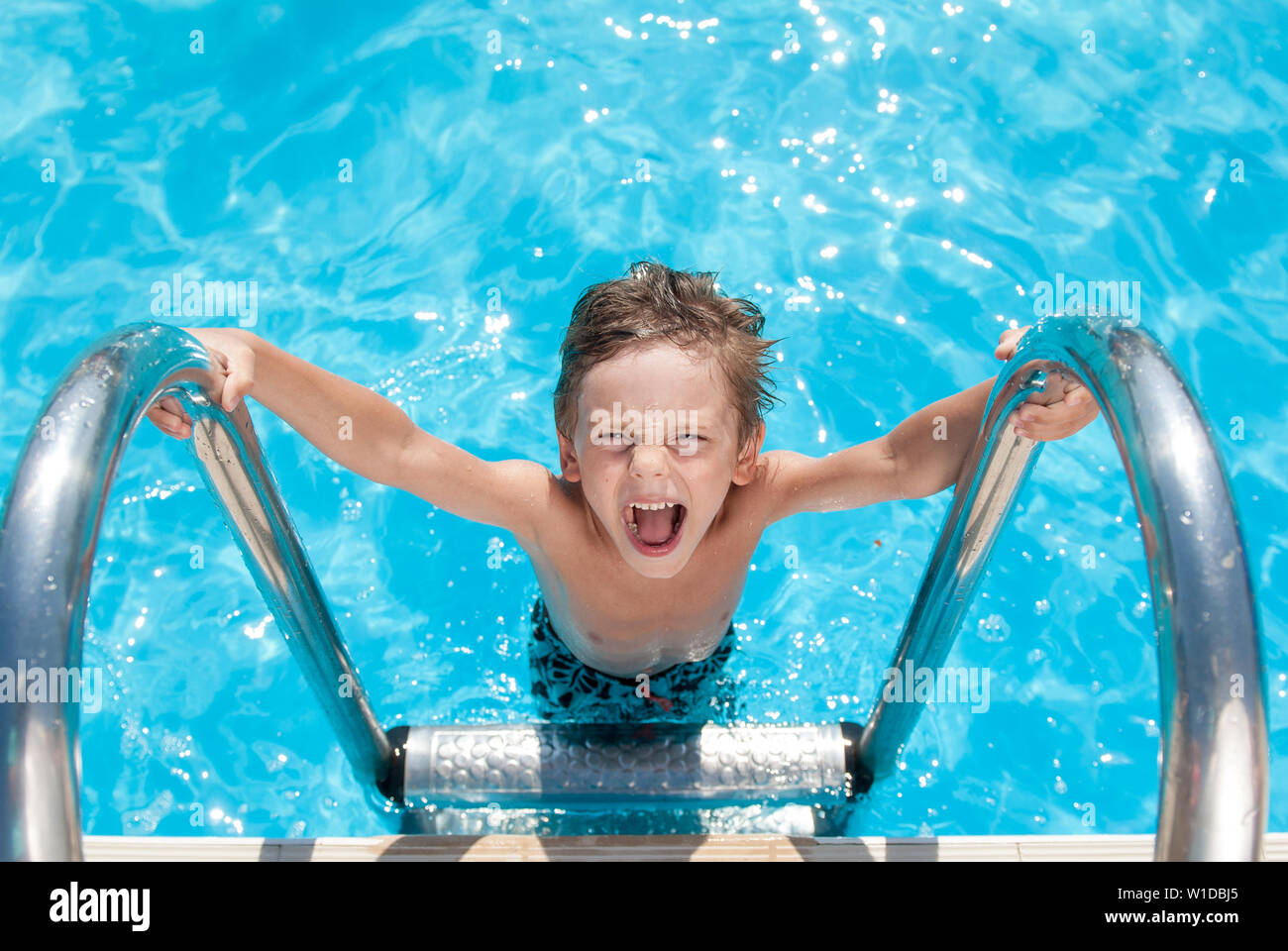 happy beautiful small shouting kid with wet hair standing on chrome handrail in fresh water swimming turquoise outdoors pool in sunny summer holiday d Stock Photo