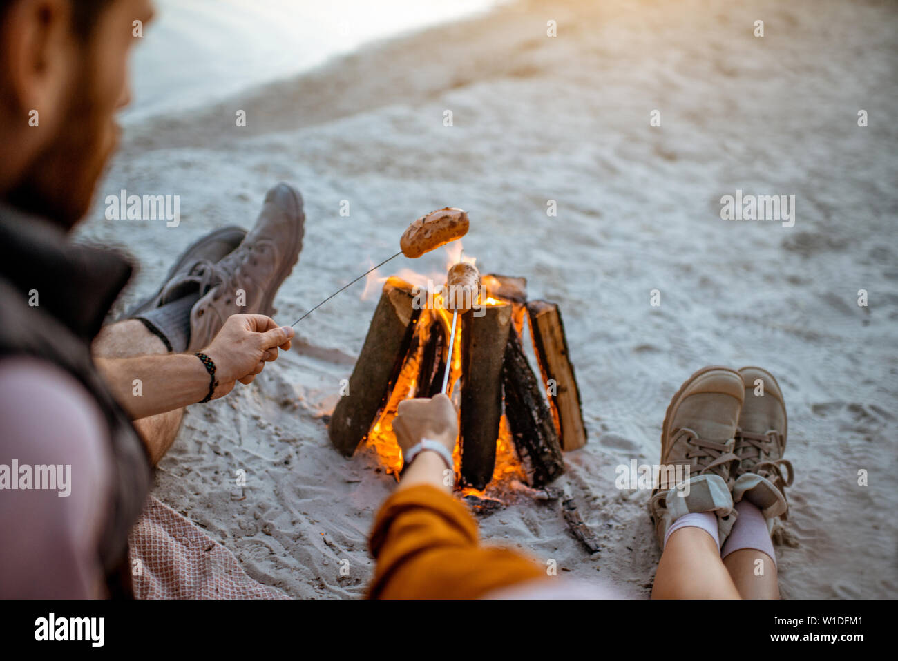 Couple cooking sausages at the fireplace, having a picnic on the beach in the evening, rear view Stock Photo