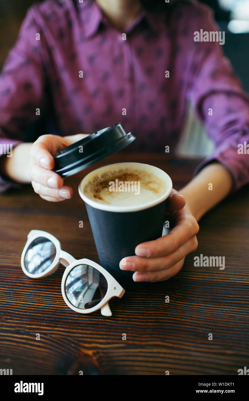 Female hands holding a cup of cappuccino with thick foam in black takeaway paper cup. Coffee take away. Stock Photo