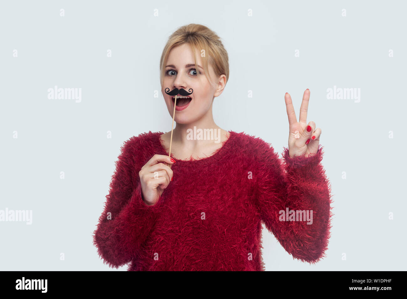 Beautiful young woman in red blouse standing, holding manlike mustache sticker, showing peace sign with two fingers and opened mouth, looking at camer Stock Photo