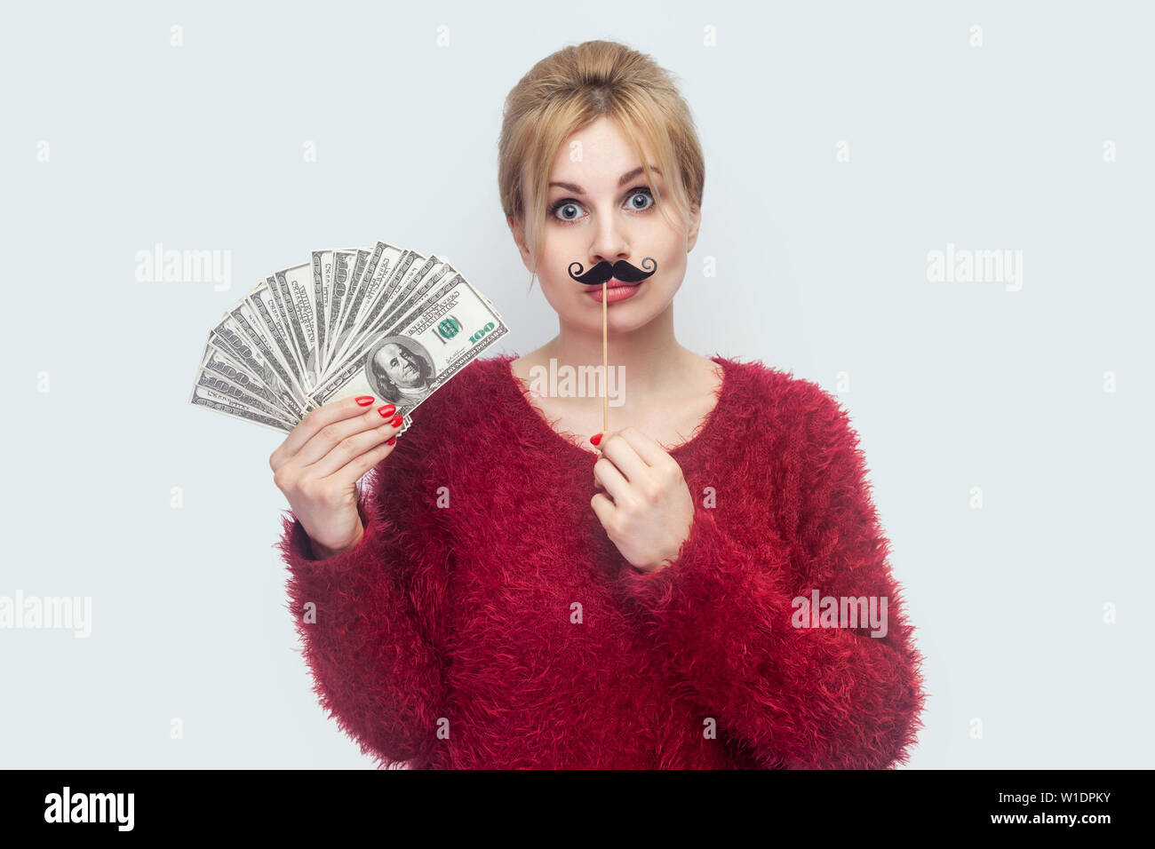 Beautiful young woman in red blouse standing, demonstrate fan of money, holding manlike mustache sticker, looking at camera with big eyes. Indoor, stu Stock Photo