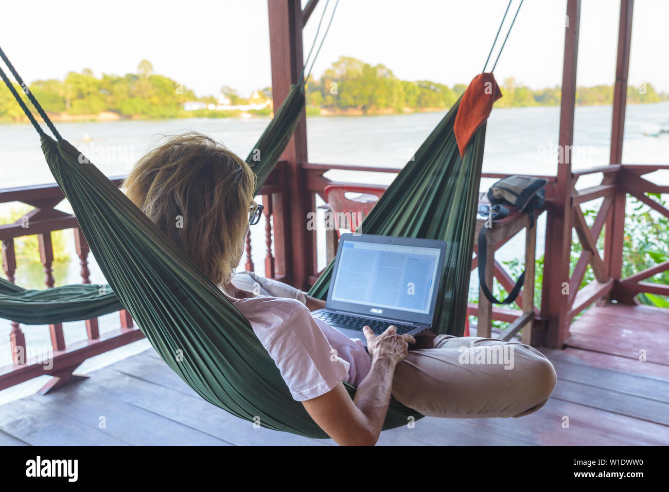 Woman working with laptop on hammock hanging in tourist resort on the Mekong River, Laos. Concept of millenials working around the world and digital n Stock Photo