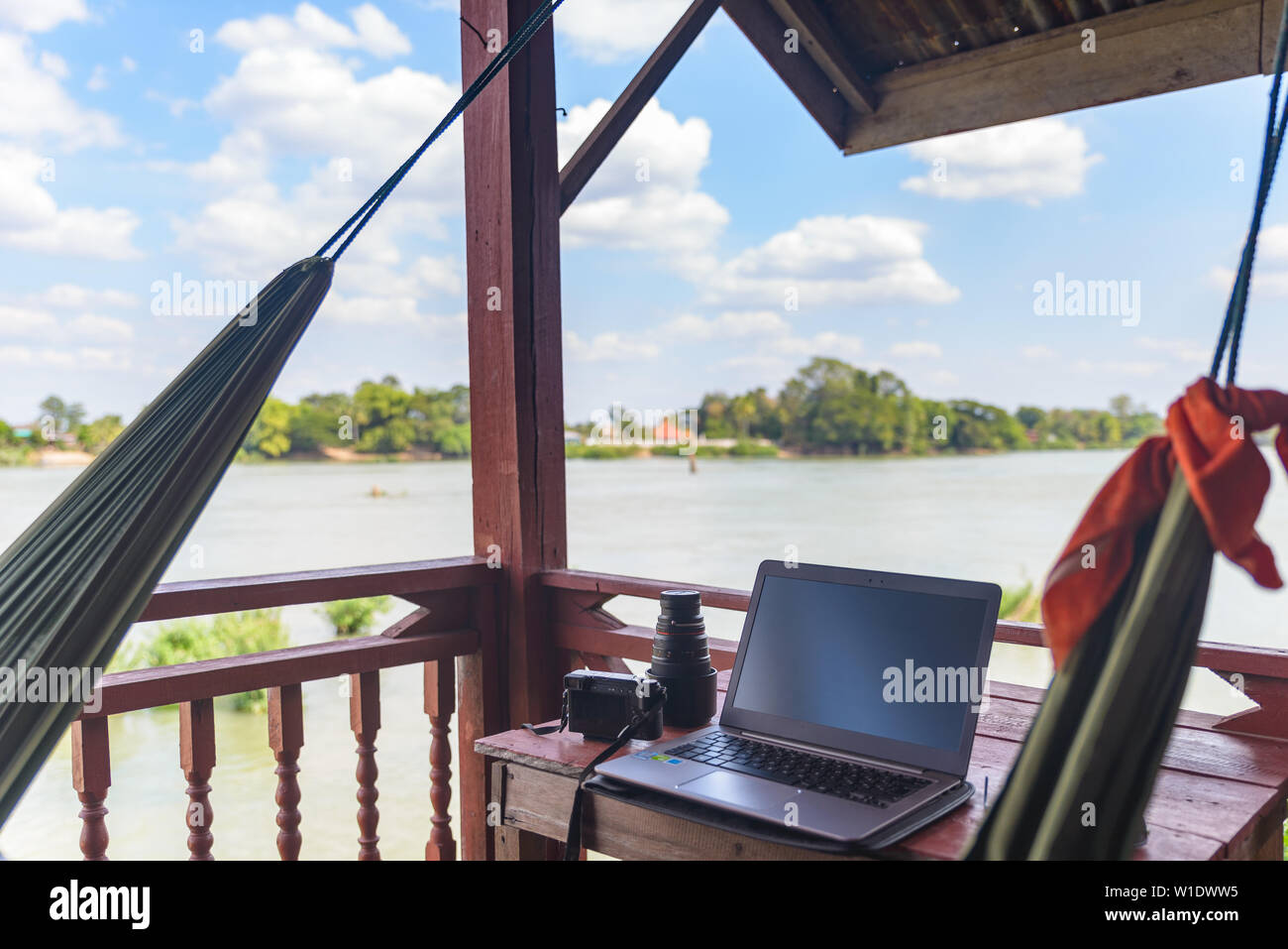 Working with laptop on wooden balcony in tourist resort with hanging hammock on the Mekong River, Laos. Concept of millenials working around the world Stock Photo