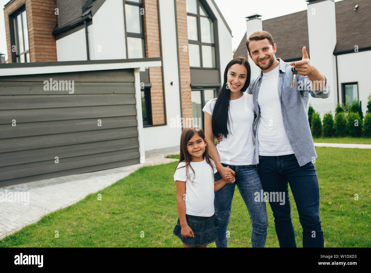 Happy heteroseksual family standing near their new house. Man holding keys from new apartment. Stock Photo