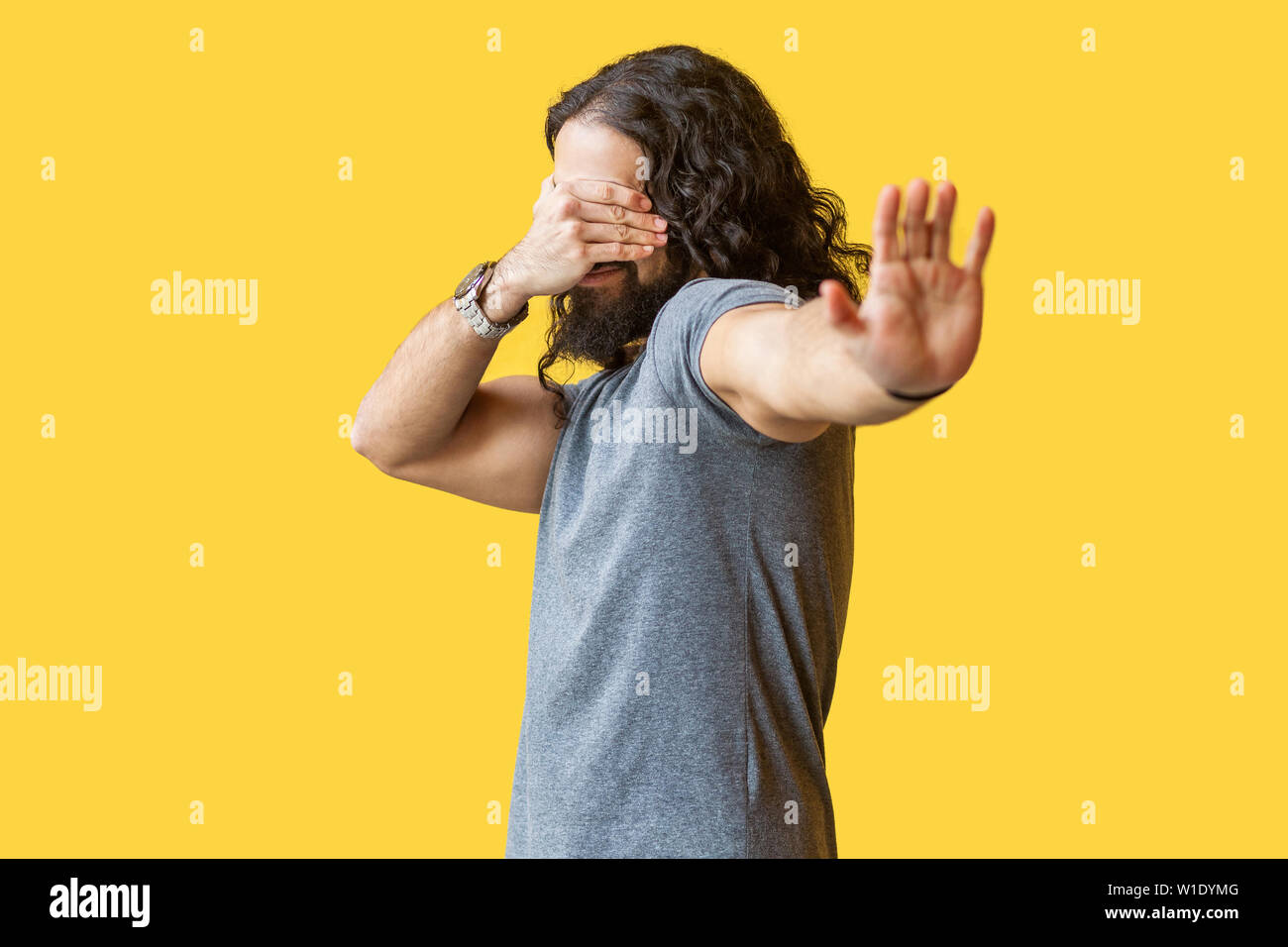 I don't want to look at this. Portrait of afraid or shy young man with long curly hair in grey tshirt closing his eyes and showing stop hand gesture. Stock Photo