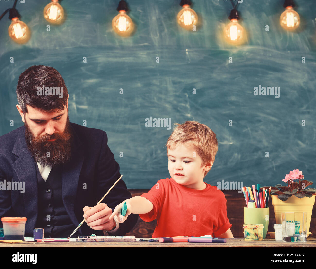 Teacher with beard, father teaches little son to draw in classroom, chalkboard on background. Child and teacher on busy face painting, drawing. Art lesson concept. Talented artist spend time with son. Stock Photo