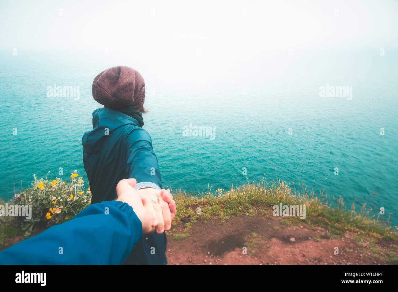 Follow me. Girl taking hand of her boyfriend walking to the cliff edge overlooking the sea on a moody and rainy day. Outdoors travel concept Stock Photo