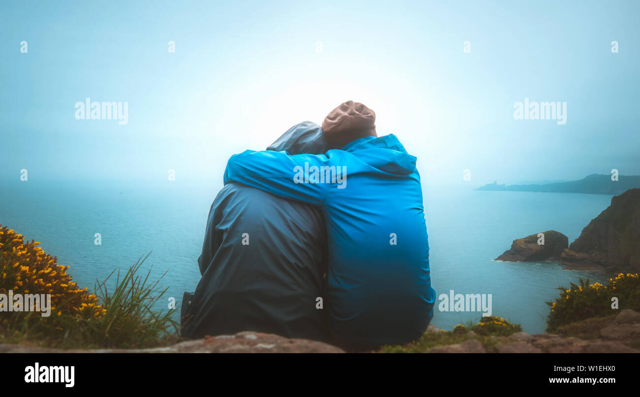 Back view or rear view of two young adults lovers sitting on the edge of a cliff watching the sea on a rainy and moody day. Partnership, outdoors, tra Stock Photo