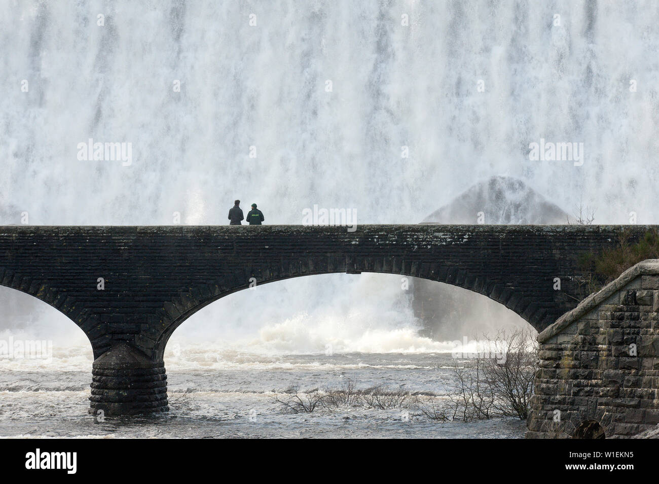 Visitors walk across a bridge as water cascades over the Caban-coch dam, at Elan Valley village near Rhayader in Powys, Wales, United Kingdom, Europe Stock Photo
