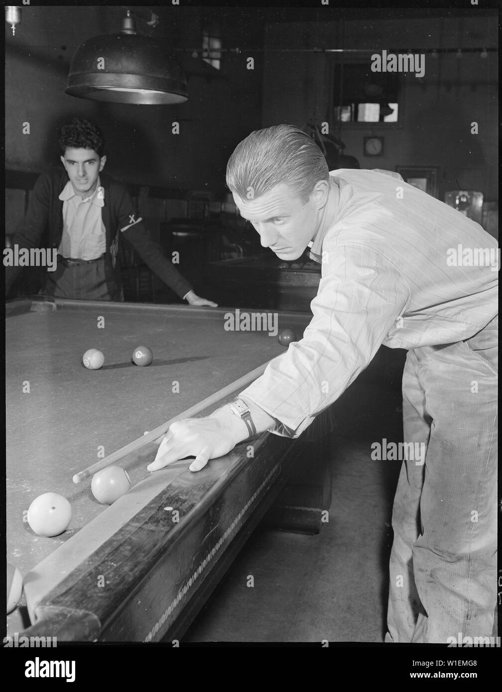 Hayward, California. Pool Recreation. Ten minutes to four by the wrist watch of this high school boy with the cue. In contrast to the gloomy, ill-lit interior of the pool hall, it was a sunny Saturday afternoon outdoors. These two boys were part of a group that had been loafing around the pool hall since about 10:00 that morning. They planned to drift together again the next day and spend their Sunday in the same idle manner Stock Photo
