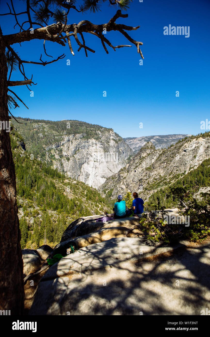 Tourists hikers sitting on the edge of a cliff at the shade of a tree in John Muir Trail, in summer with clear blue sky, Yosemite National Park Stock Photo