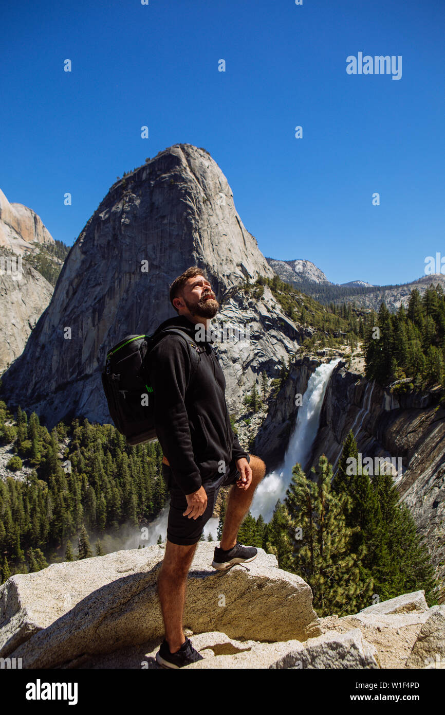 Tourist man with backpack posing in John Muir Trail with Nevada Fall and Liberty Cap, in summer with clear blue sky, Yosemite National Park Stock Photo