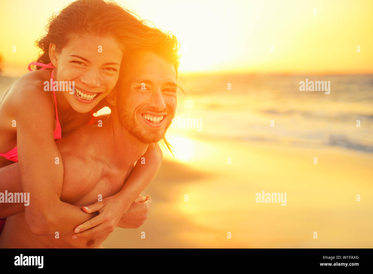 Lovers couple in love having fun dating on beach portrait. Beautiful healthy young adults girlfriend piggybacking on boyfriend hugging happy. Multiracial dating or healthy relationship concept. Stock Photo