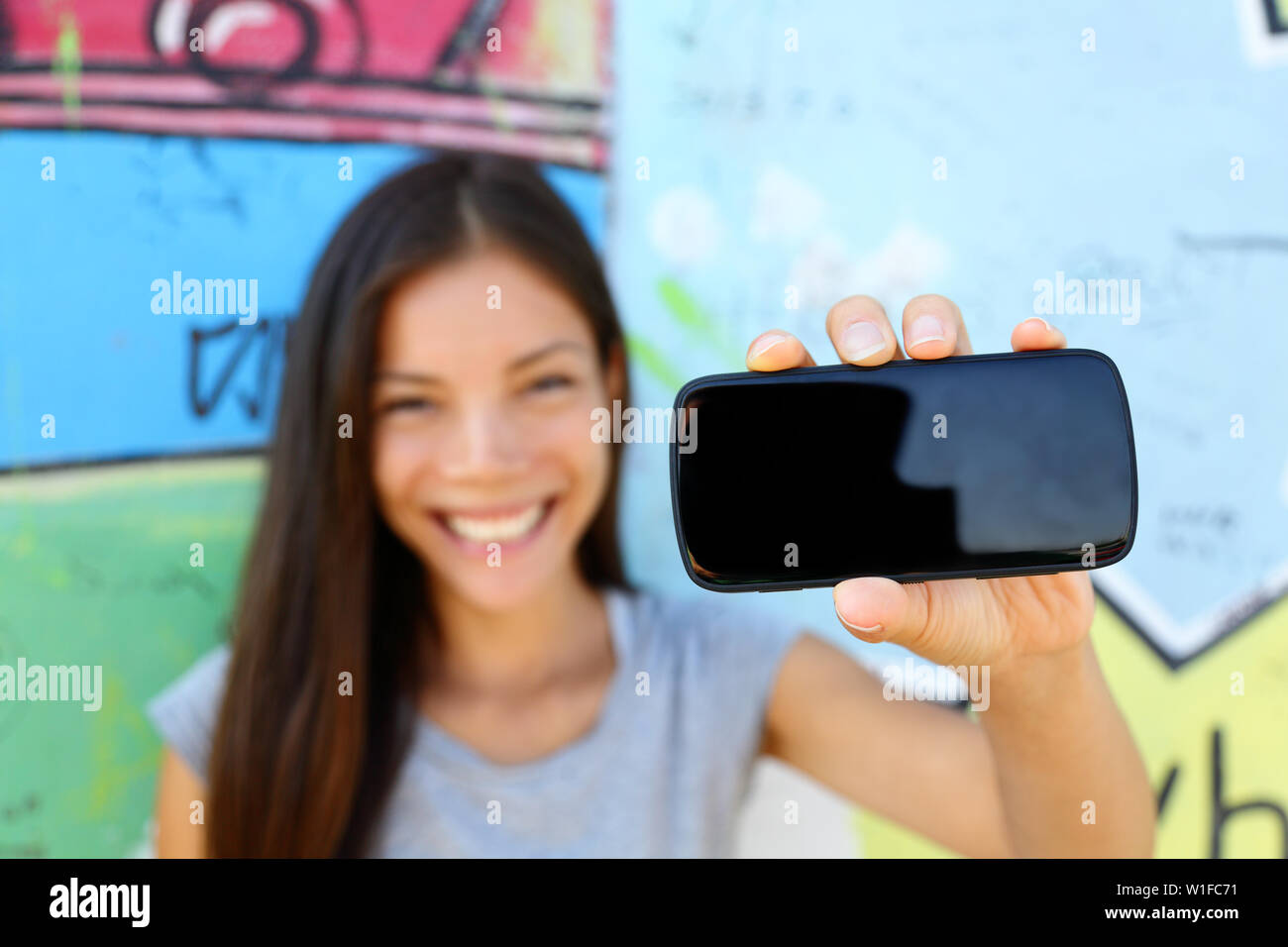 Smartphone - young ethnic woman showing screen for copyspace for text or photo. Asian young adult holding phone with urban graffiti background at Berlin wall, Germany. Stock Photo