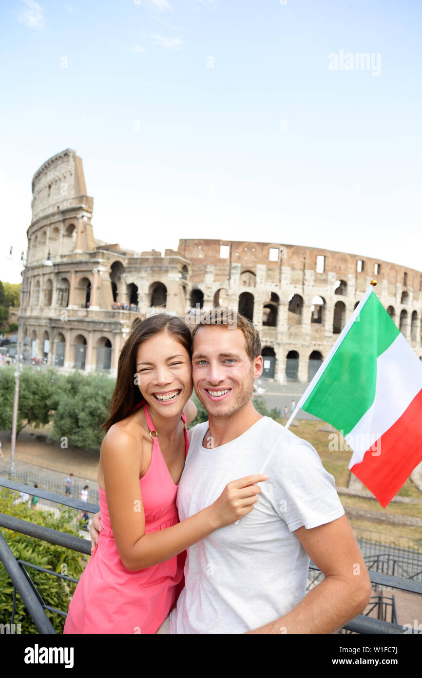 Happy travel - couple tourists in front of Coliseum, Rome, Italy. Cheerful young Asian and Caucasian adults posing with Italian flag during their European holiday vacations in Europe. Stock Photo