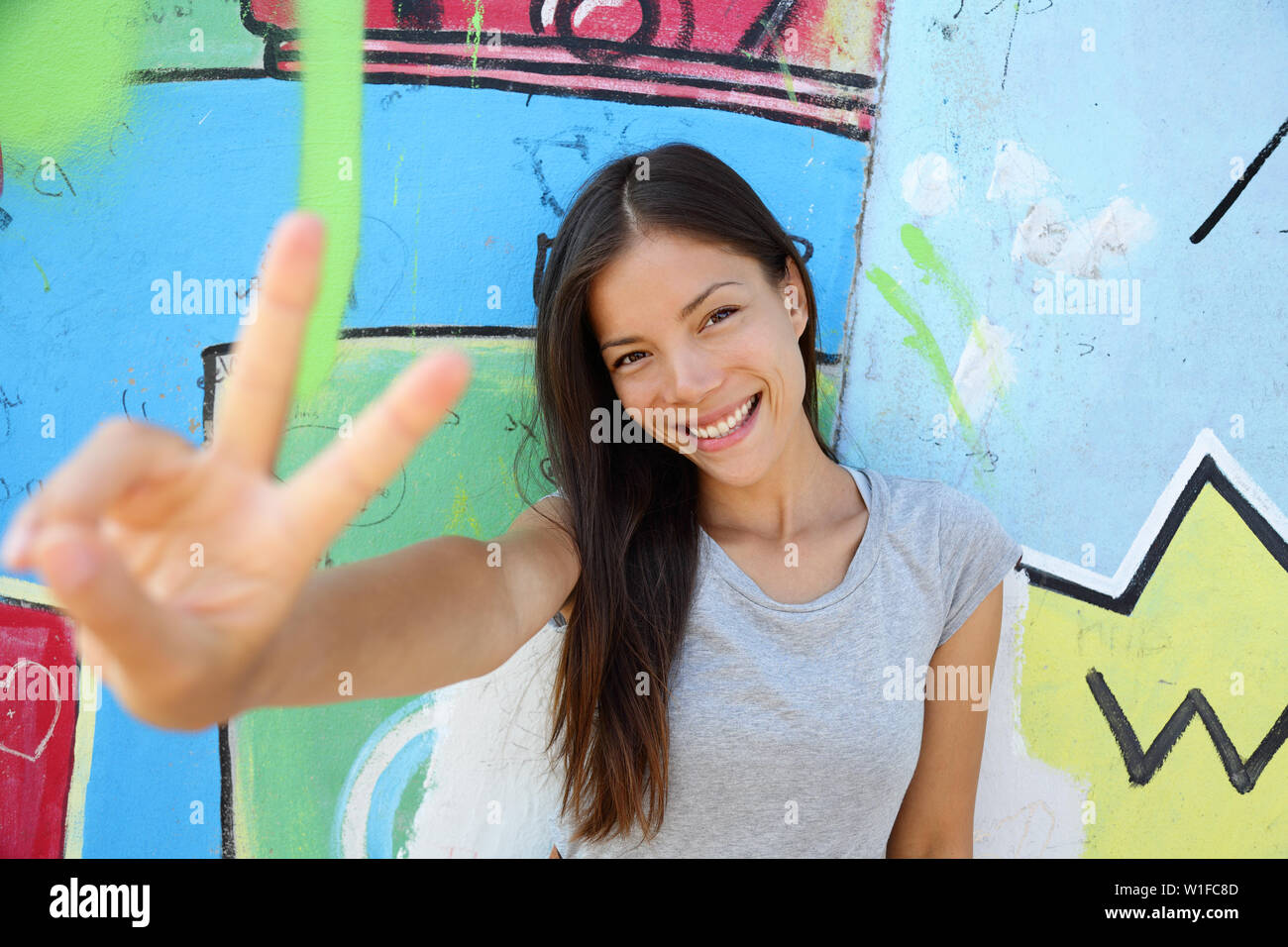 Urban young girl showing v peace sign in city. Cool Asian woman leaning on graffiti background at the Berlin wall, Germany. Modern portrait. Stock Photo