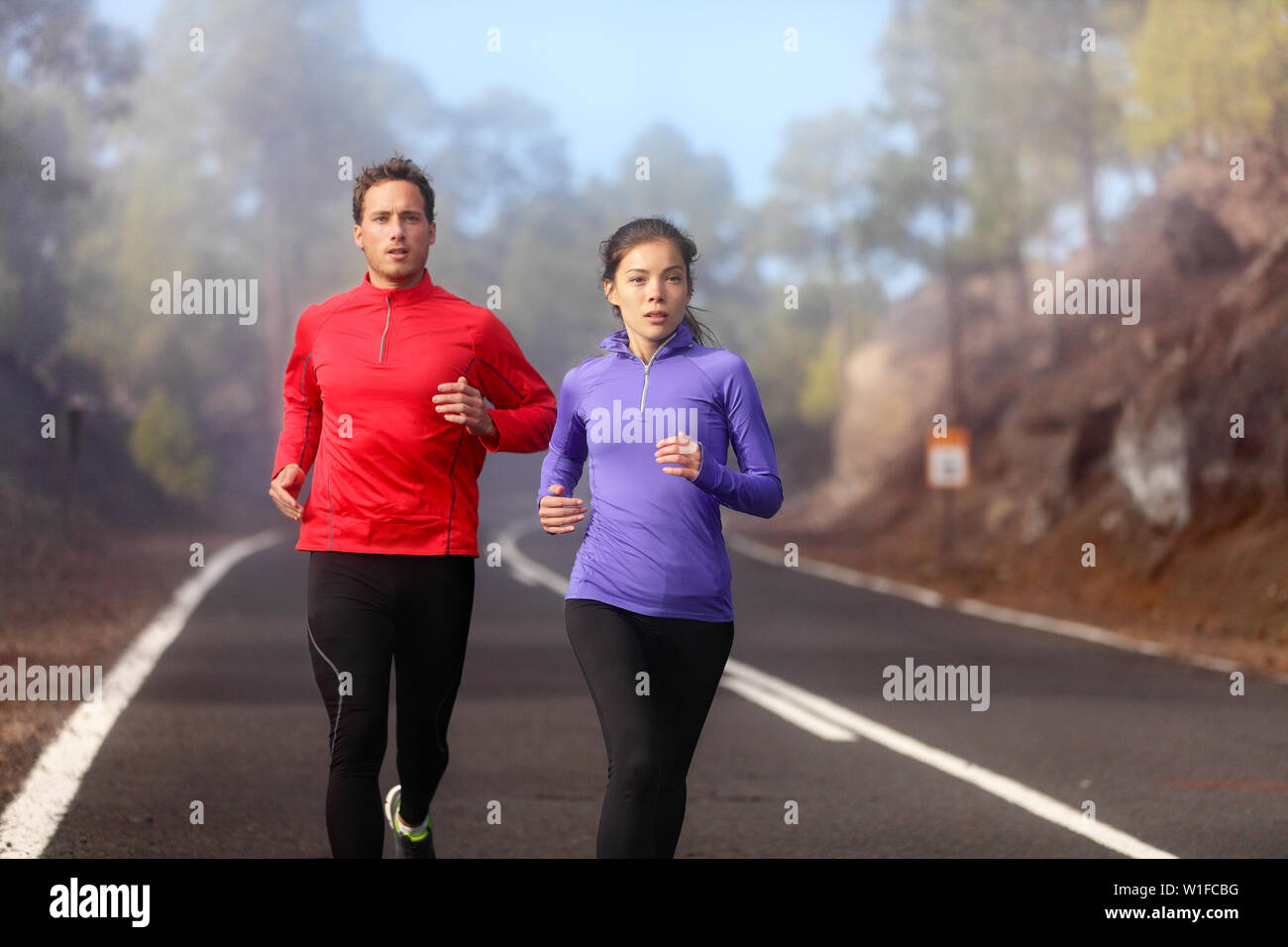 People running - athlete runners training jogging in cloudy and cold weather. Exercising runner couple working out living healthy lifestyle training for marathon together on mountain road. Stock Photo