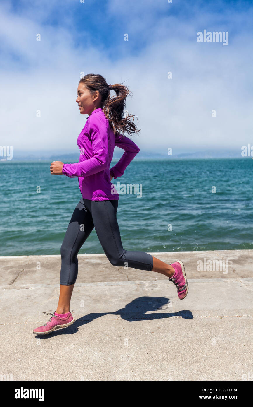 Woman running on waterfront harbour pier in San Francisco, California, USA.  Mixed race fit fitness sport model in spring / autumn running clothing  outfit. Smiling happy female athlete runner training Stock Photo -