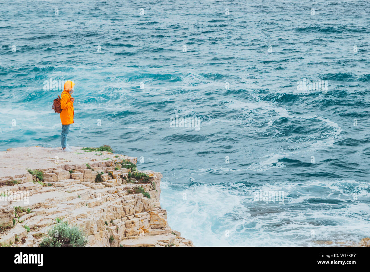 woman in yellow raincoat with backpack standing on the cliff looking at ...