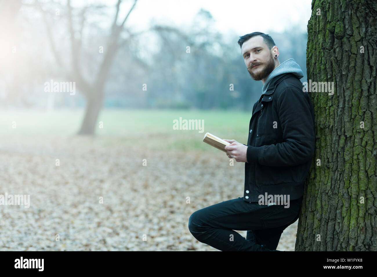 A smiling handsome young hipster man leaning against a tree and reading a book in the park. Stock Photo