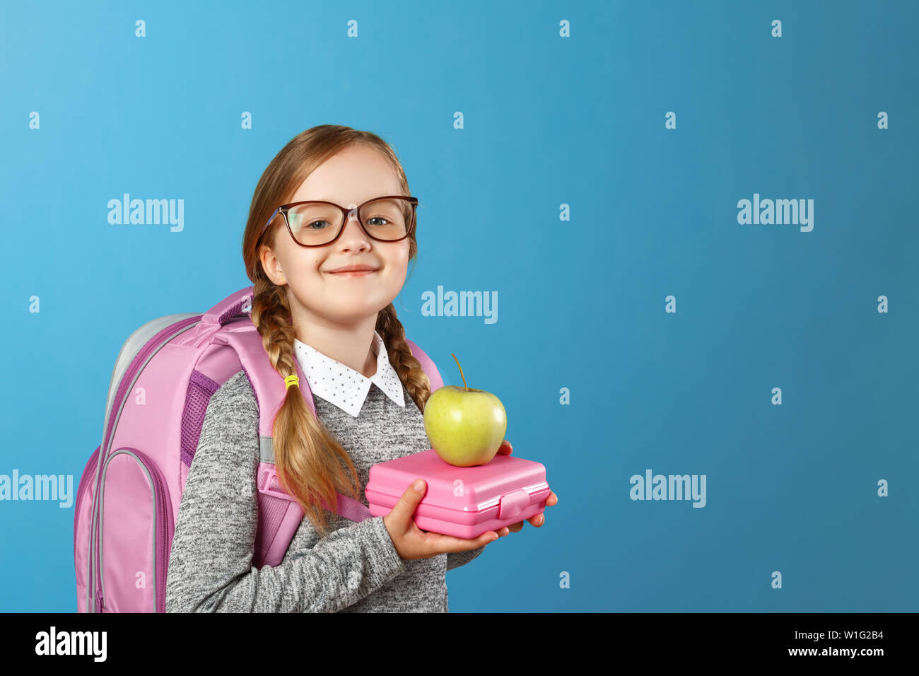 Portrait of a little girl schoolgirl in glasses with a backpack and lunch boxing on a blue background. Back to school. The concept of education. Copy Stock Photo