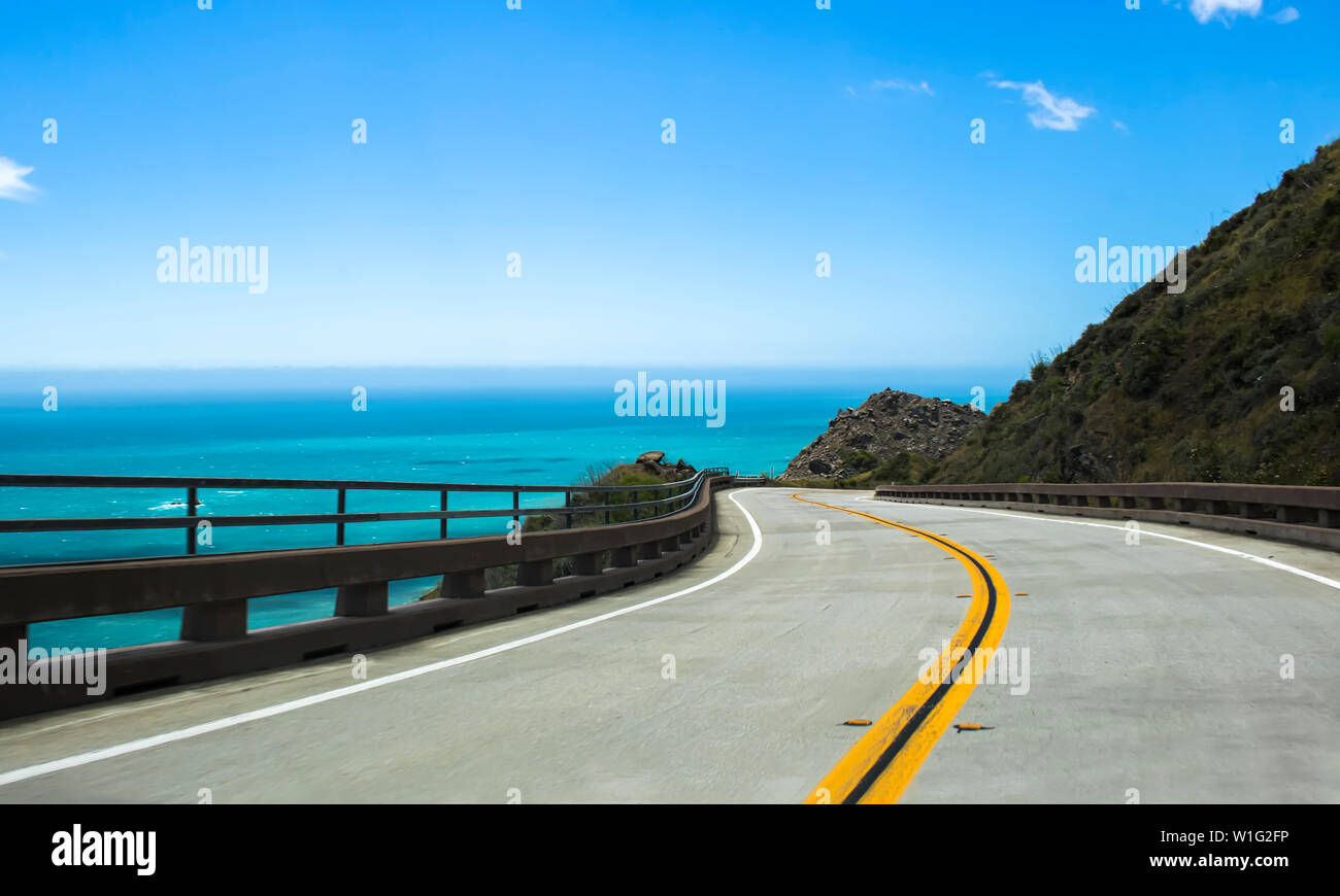 Road leads along coast of California on Highway 1 with curving pavement and guard rails over bright blue water. Stock Photo