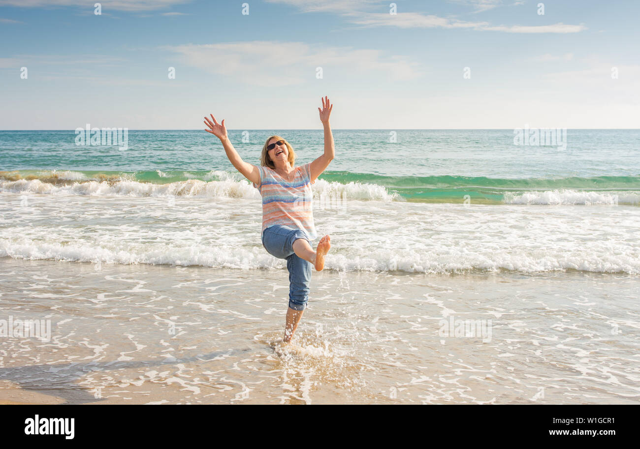 Senior woman enjoying retirement life feeling happy and healthy playing with the water and having fun in the beach in a sunny beautiful day. In Active Stock Photo