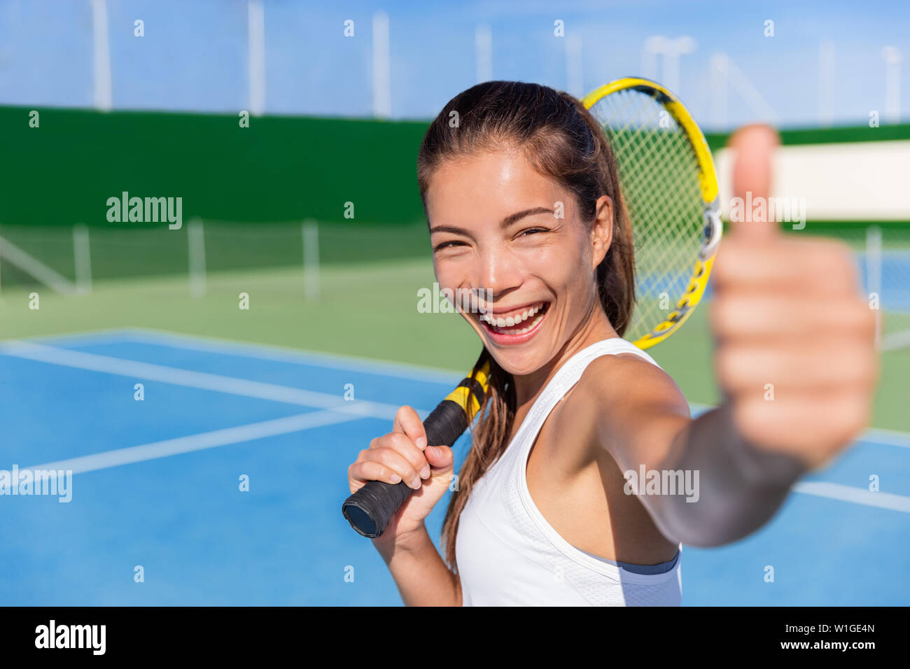 Happy Asian tennis player girl showing thumbs up hand sign after game fun holding racket on outdoor blue hard court for summer class. Sports ethnic young woman smiling of satisfaction. Stock Photo