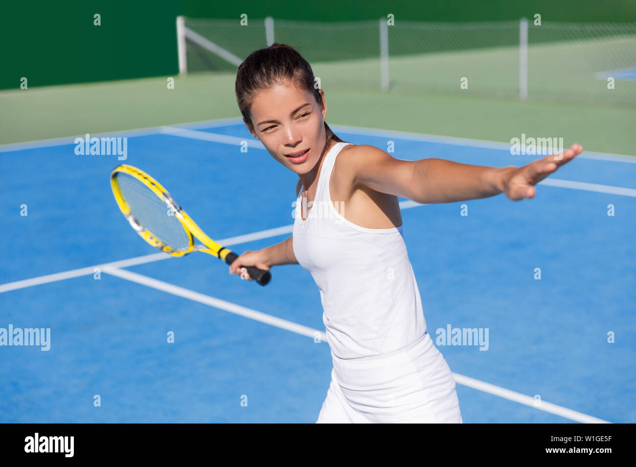 Asian tennis player woman playing hitting forehand in white dress outfit on blue hard court outdoor in summer holding racket. Female athlete determination and concentration concept. Stock Photo
