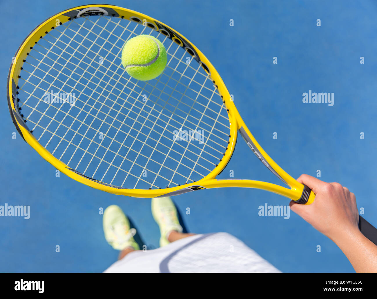 Tennis player holding yellow ball on racket grid. Sports female athlete taking a feet selfie showing running shoes on blue hard court. POV closeup of equipment, neon yellow fashion footwear. Stock Photo