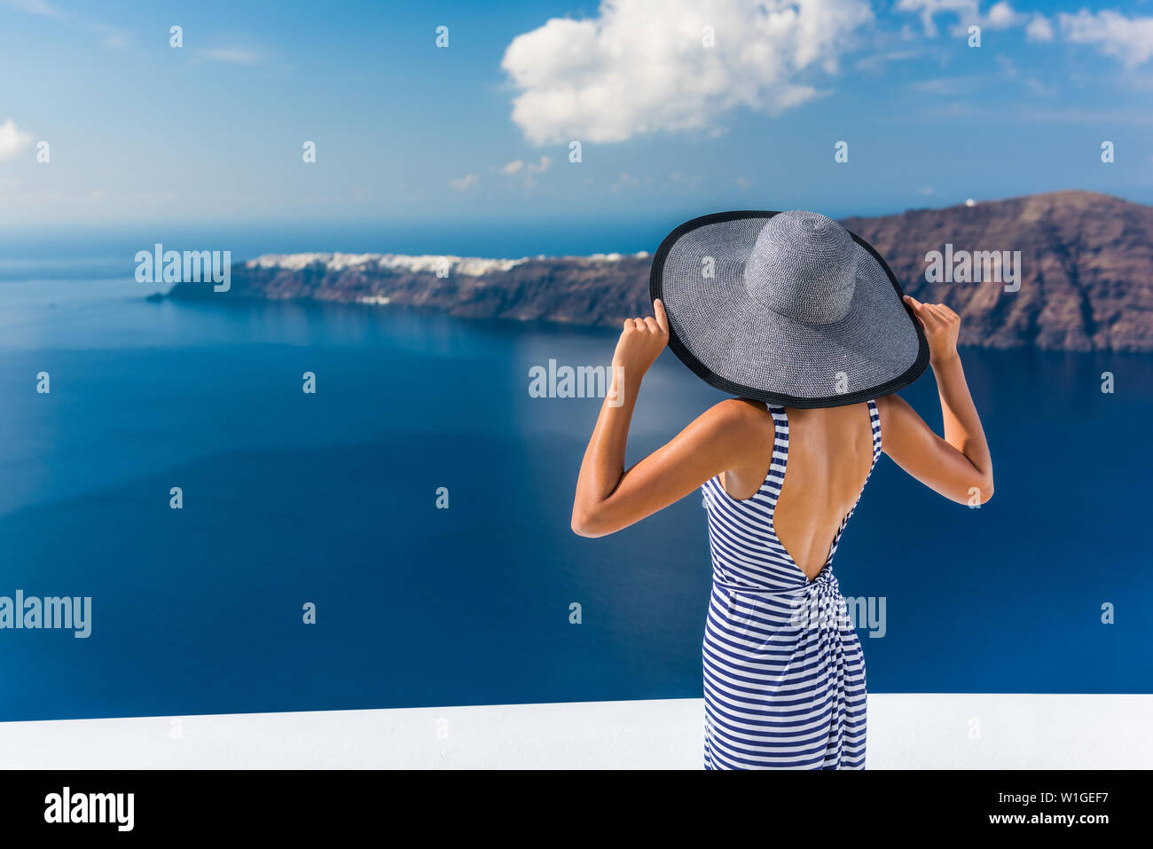 Europe summer vacation travel destination luxury living woman looking at view of Mediterranean Sea and Santorini island Oia village. Elegant tourist lady in fashion back dress and floppy sun hat. Stock Photo