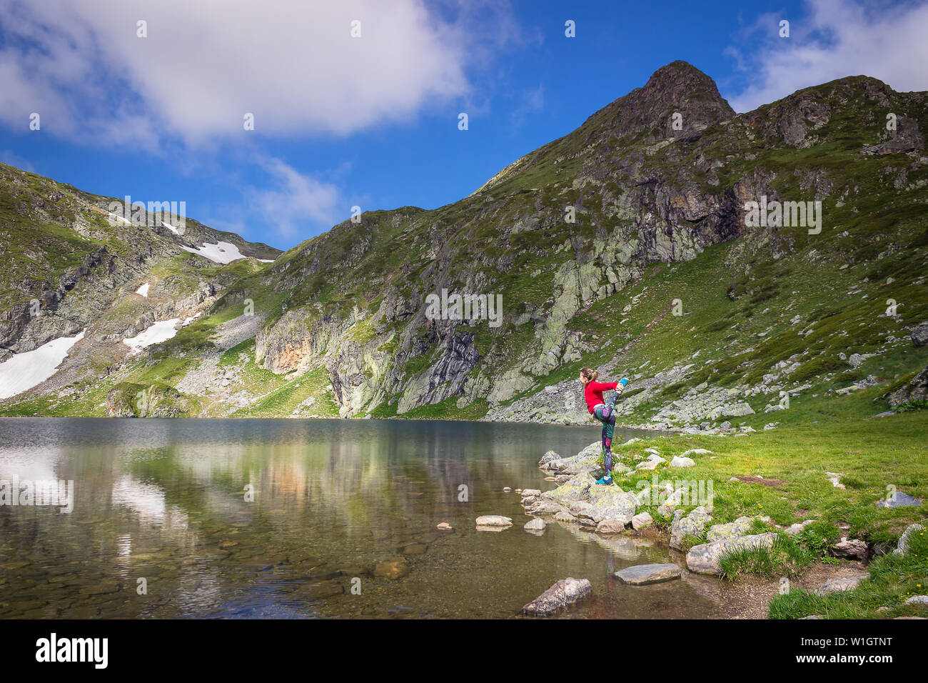 Girl with red jacket and colorful tights standing on one leg at the edge of beautiful Kidney lake on Rila mountain and epic, rocky peaks Stock Photo