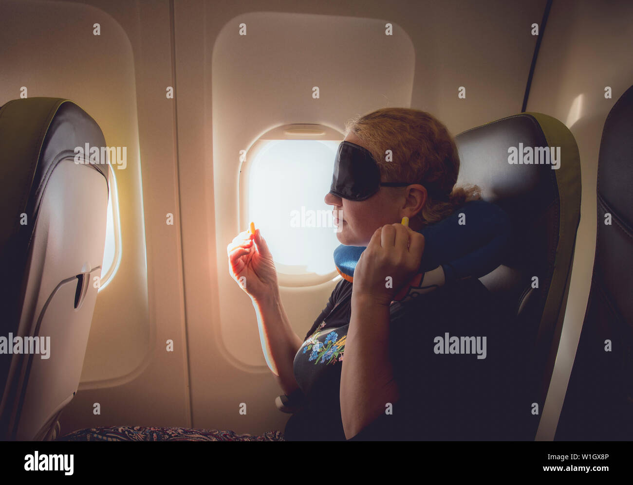 33 year old woman using traveling pillow, sleeping mask and ear plugs in plane cabin on passenger seat, fly with airplane, comfortable cozy traveling Stock Photo