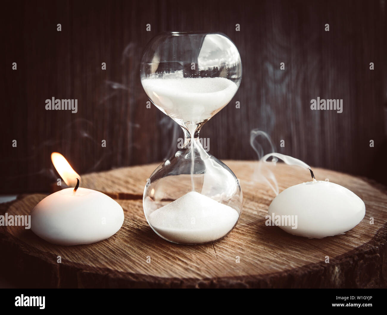 Time running out, life and death concept. One candle burning with flame as living other is blown away as dead and hourglass measures lifetime. Stock Photo