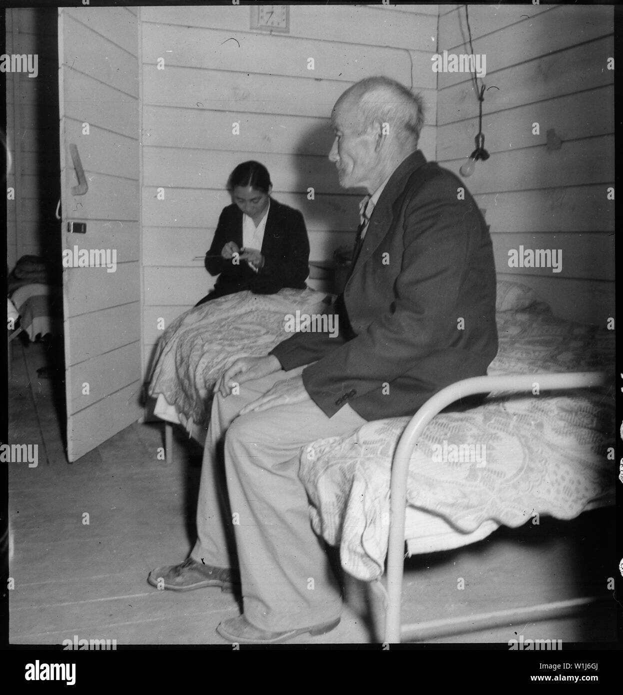 San Bruno, California. Old Mr. Konda in barrack apartment, after supper. He lives here with his tw . . .; Scope and content:  The full caption for this photograph reads: San Bruno, California. Old Mr. Konda in barrack apartment, after supper. He lives here with his two sons, his married daughter and her husband. They share two small rooms together. His daughter is seen behind him, knitting. He has been a truck farmer and raised his family who are also farmers, in Centerville, Alameda County where his children were born. Stock Photo