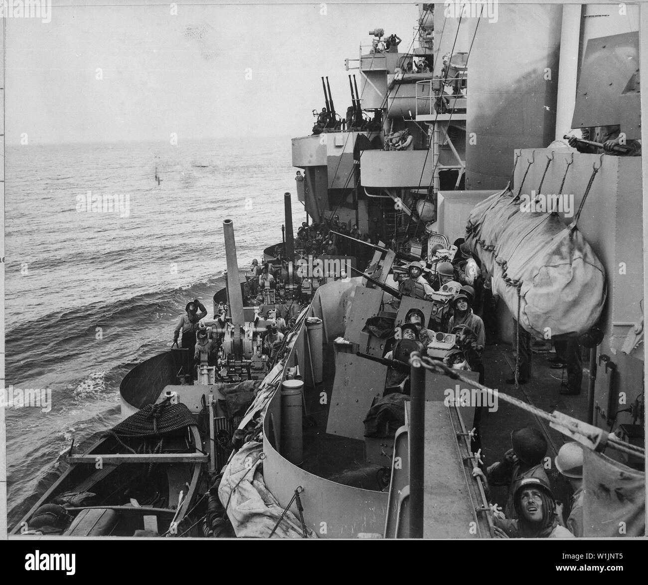 The gun crews of a Navy cruiser covering American landing on the island of Mindoro, December 15, 1944, scan the skies in an effort to identify a plane overhead. Two 5 inch (127mm) guns are ready while inboard 20mm anti-aircraft crews are ready to act.; General notes:  The cruiser is USS Phoenix (CL-46), identifiable by her unique sprayed camouflage Measure 32 Design 5d. Stock Photo