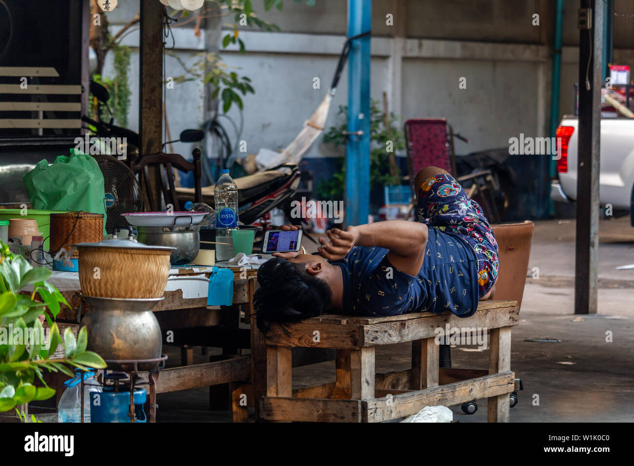 Bangkok, Thailand - April 12, 2019: Man lying on a bench, smoking and listening to music on a street in central Bangkok Stock Photo