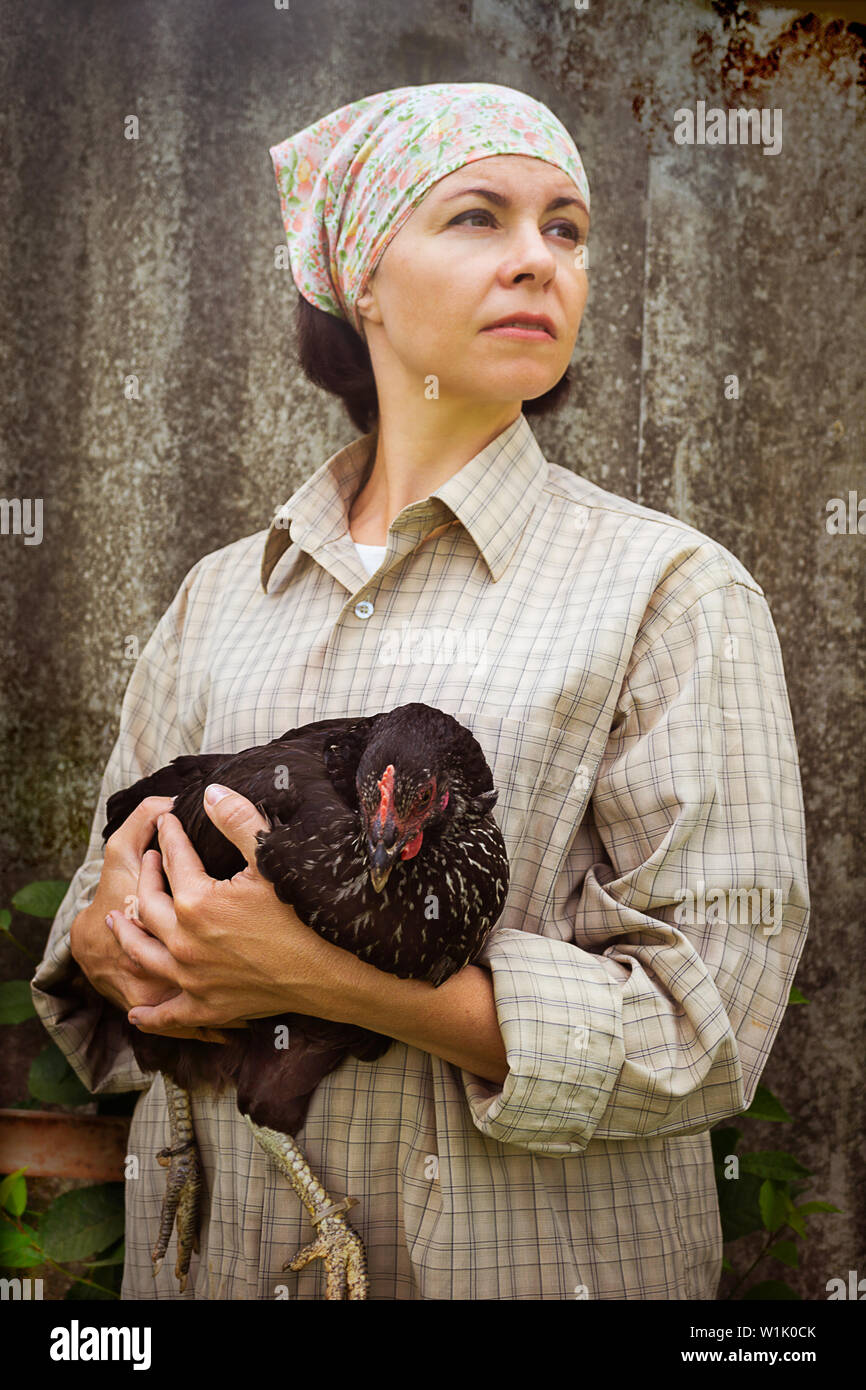 woman farmer in men's clothing, holding a black chicken in her hands in the rural scene. Stock Photo