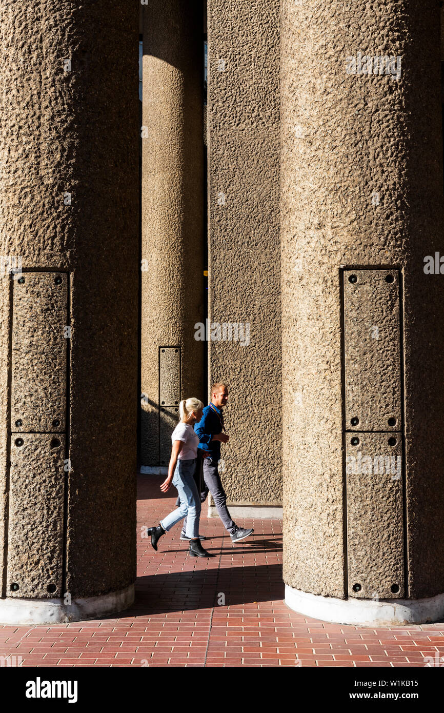 A couple walk past Brutalist concrete pillars at the Barbican Estate in London EC2, England, UK. Stock Photo