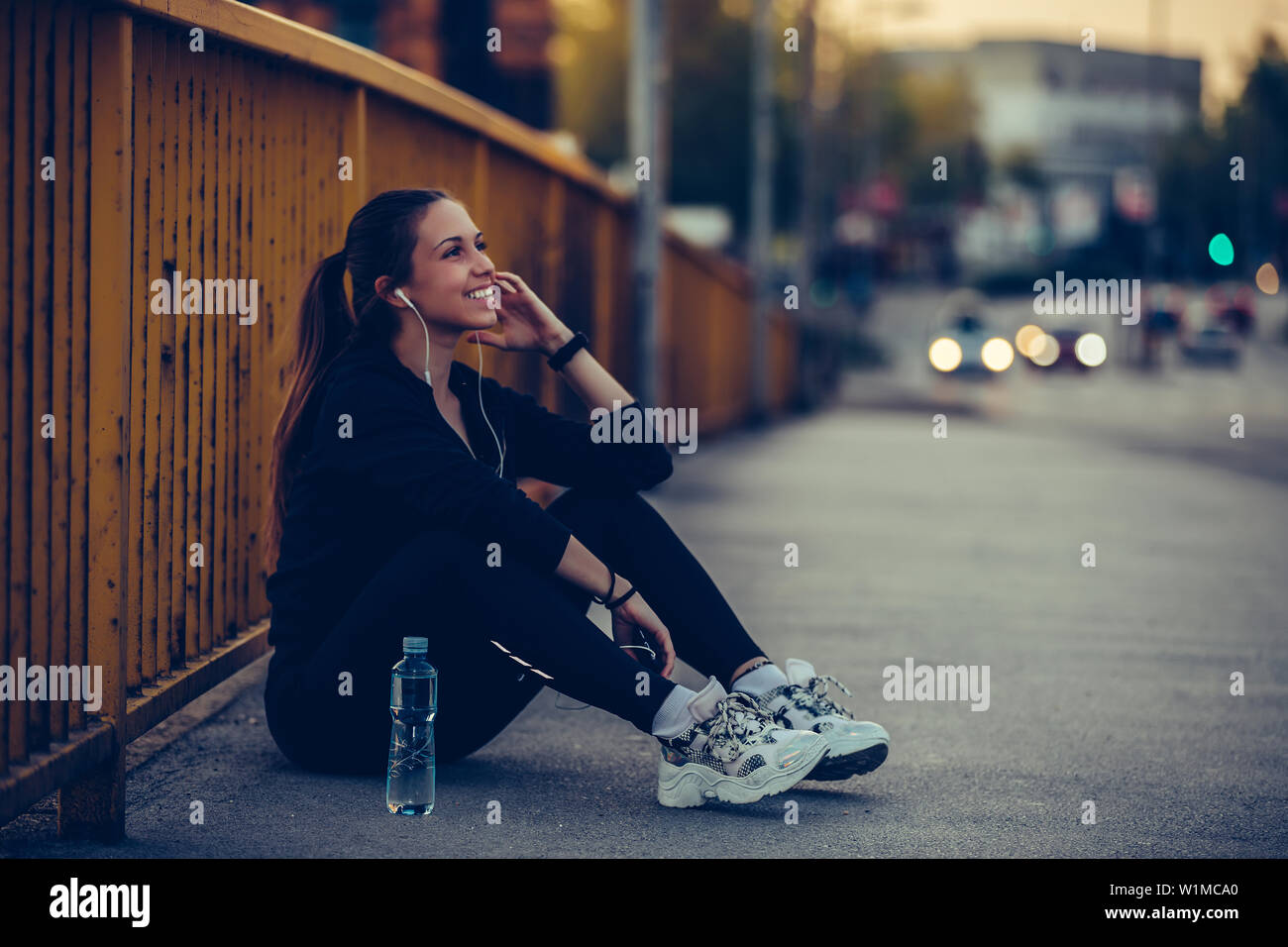 Young woman in black sports outfit listening to music and resting after running on the bridge in the city, at night Stock Photo