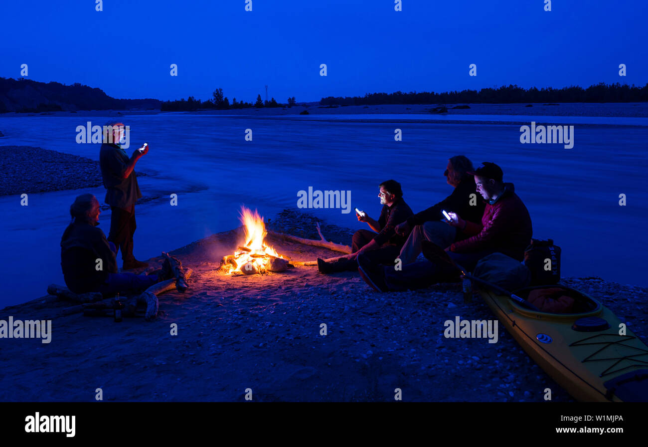 Group of paddlers with smart phones at camp fire, Tagliamento river, Friuli, Italy Stock Photo
