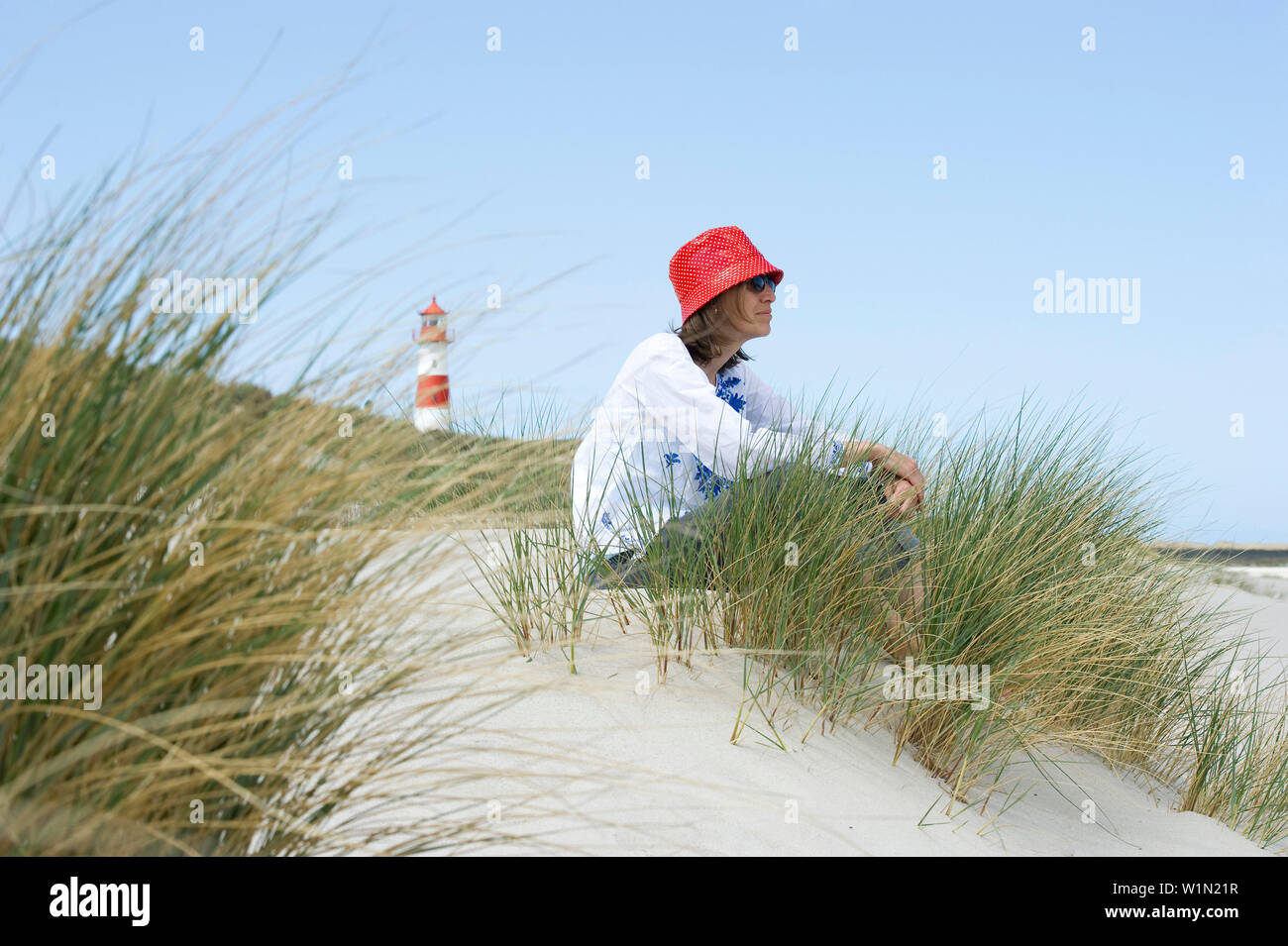 Woman wearing a red hut sitting at beach, List-Ost lighthouse in background, Ellenbogen, List, Sylt, Schleswig-Holstein, Germany Stock Photo