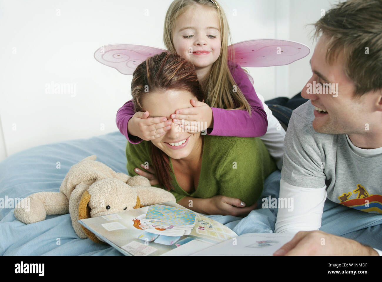 Young family lying on bed while reading a book, Munich, Germany Stock Photo
