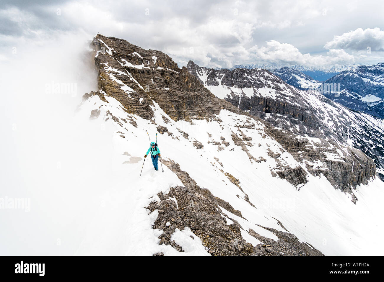 Young woman ascending a mountain ridge with skis on the backpack during wintertime, clouds moving in, Birkkarspitze, Karwendel, Tyrol, Austria Stock Photo